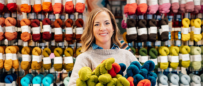 Jill, owner of North Bay Fiber, smiles as she holds a dozen skeins of yarn.